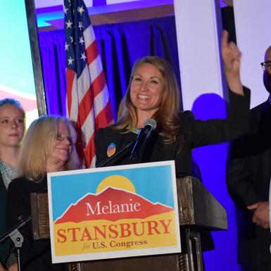 Melanie Stansbury celebrates with supporters on stage behind a podium with her campaign logo along